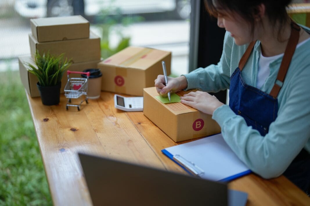 asian entrepreneur working in small start up business online shop company. woman using tape to seal box by packing product to send customers in office. elegant female packaging parcels
